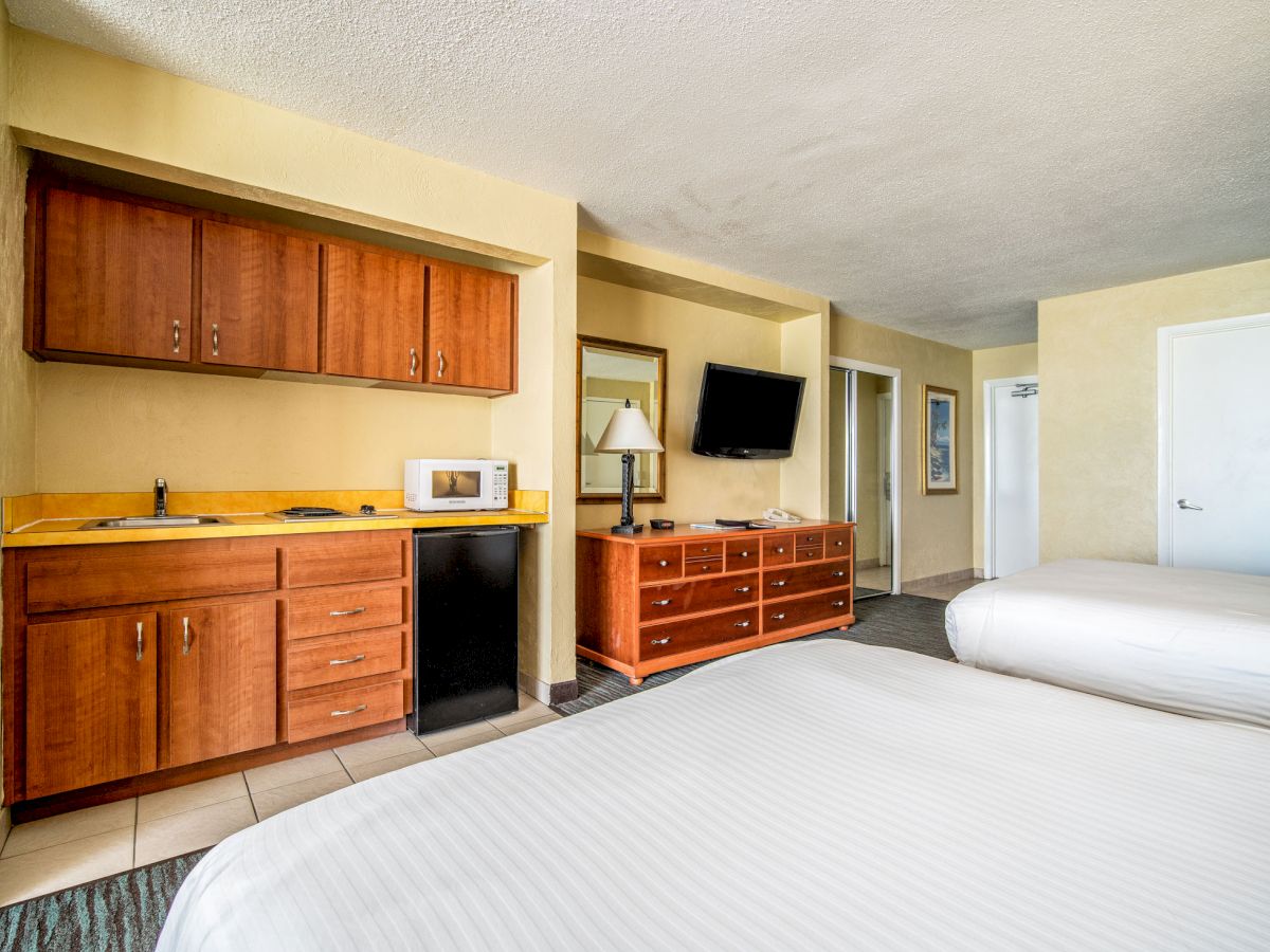 This image shows a hotel room with two beds, a TV on a dresser, and a kitchenette with a sink, microwave, and mini-fridge.