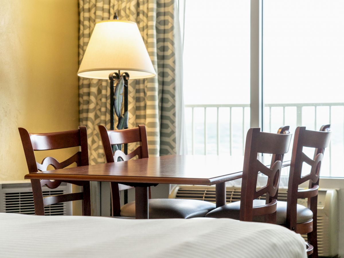 The image shows a room with a wooden table, four chairs, a lamp, curtains, and part of a bed, against a large window.