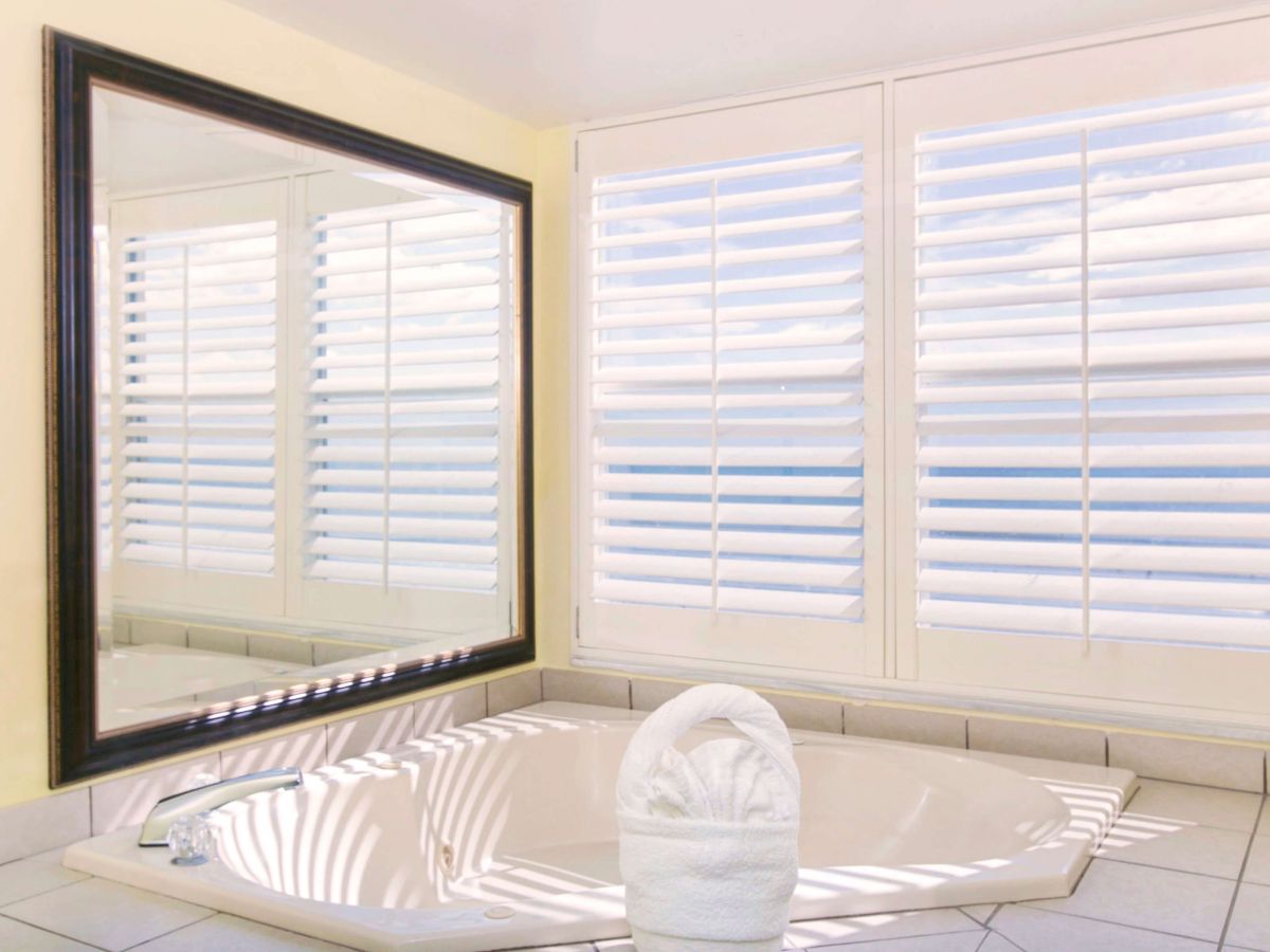 A bathroom with a large mirror, a soaking tub, white shutters on the windows, and a towel folded into a swan shape on the tub's edge.