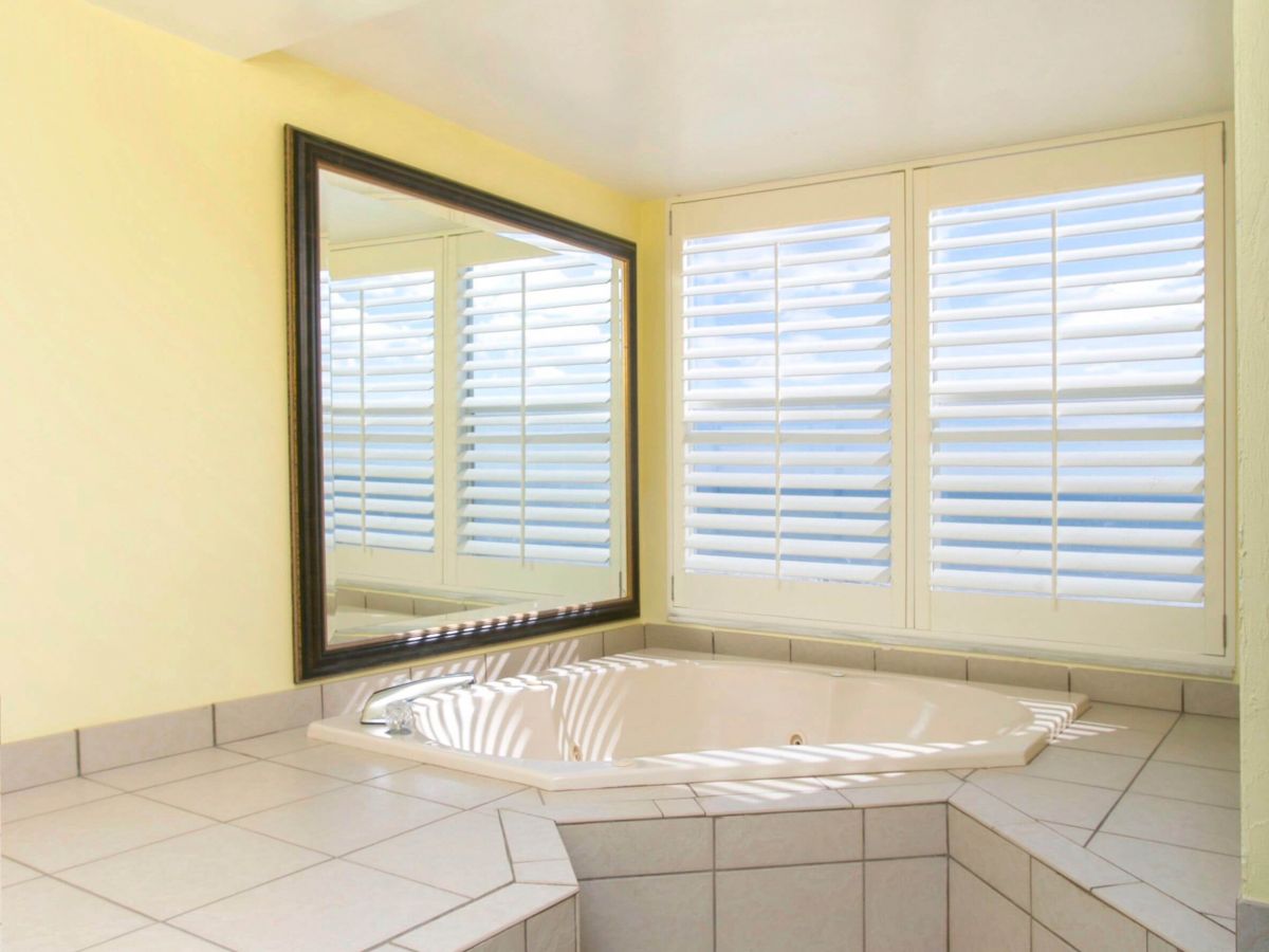 The image shows a tiled bathroom with a built-in corner bathtub, a large mirror on the wall, a ceiling light, and windows with blinds.