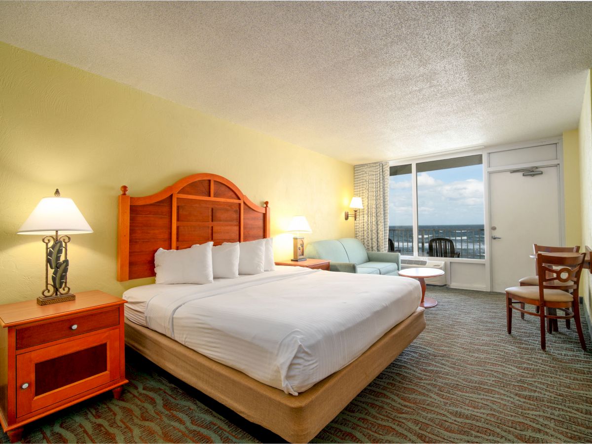 This image shows a hotel room with a king-sized bed, side table, lamp, couch, chair, small table, and balcony door with an ocean view outside.