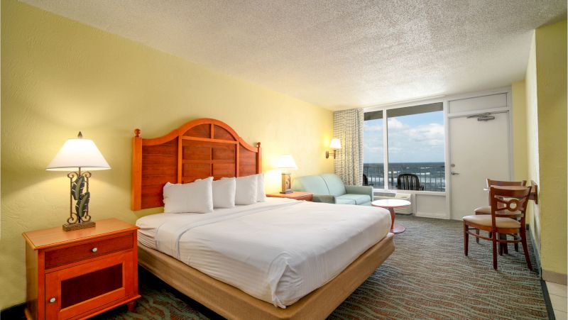 A hotel room with a bed, nightstand, lamp, sofa, table, chair, door, and window.