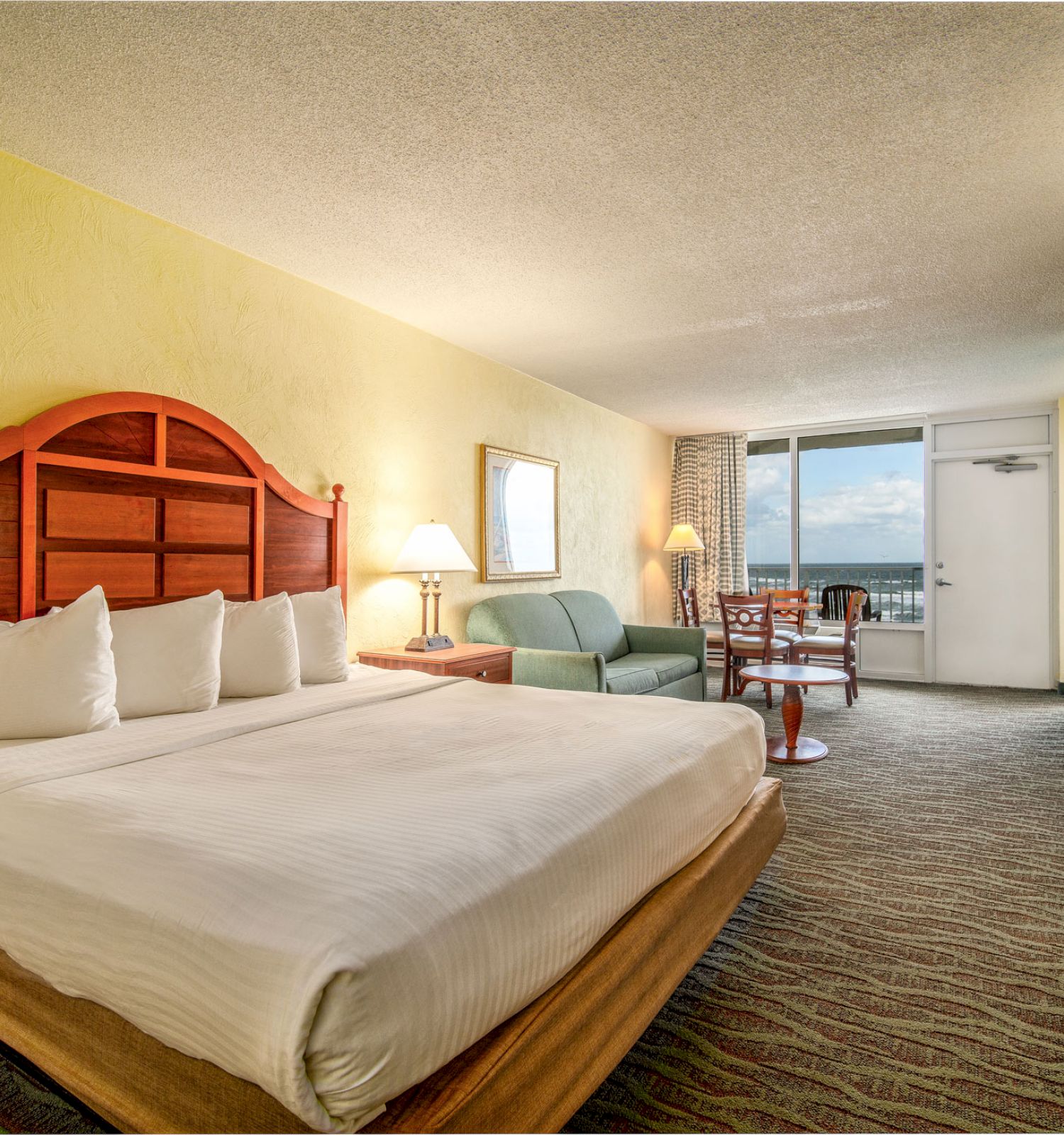 A spacious hotel room with a large bed, sitting area, table, and chairs, with a balcony overlooking the ocean visible through the window.