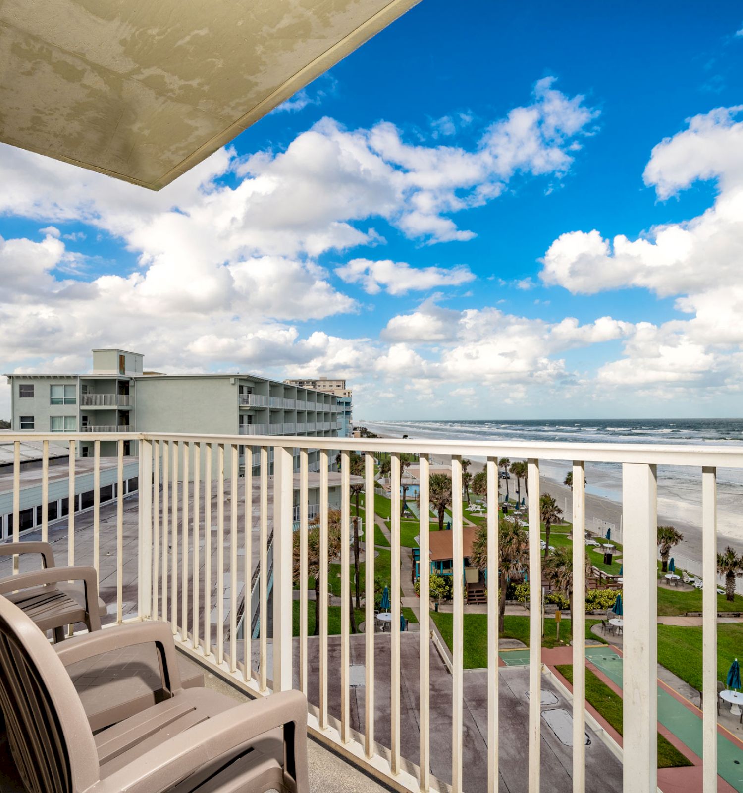 A beachfront balcony with two chairs overlooks a sandy beach and ocean waves under a partly cloudy blue sky, with nearby buildings in view.