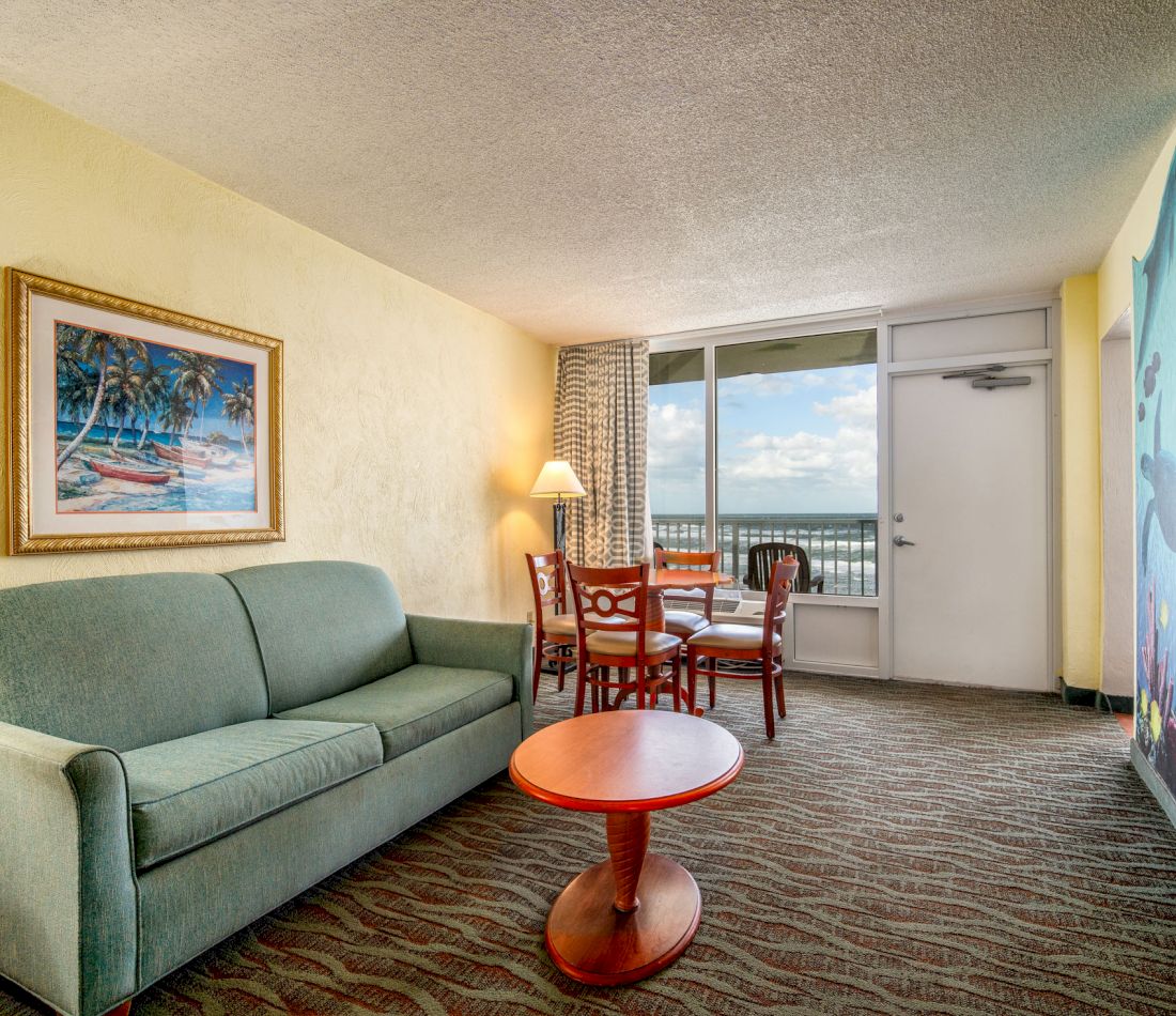 A hotel room living area with a sofa, coffee table, dining table, chairs, lamp, and artwork, overlooking the ocean.