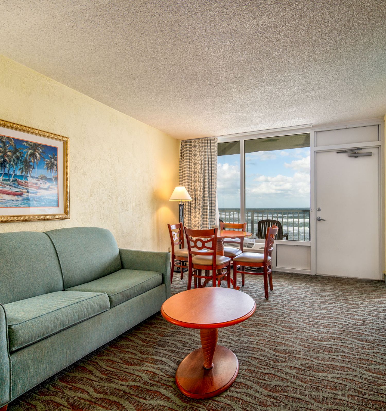 The image shows a living room featuring a green couch, a small round table, a dining set, wall art, and a door leading to a balcony with a view.