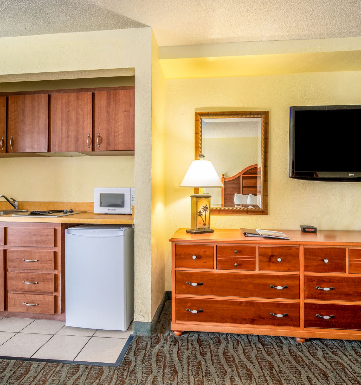 The image shows a room with a kitchenette, including a mini-fridge and microwave, and a dresser with a TV and lamp on top.