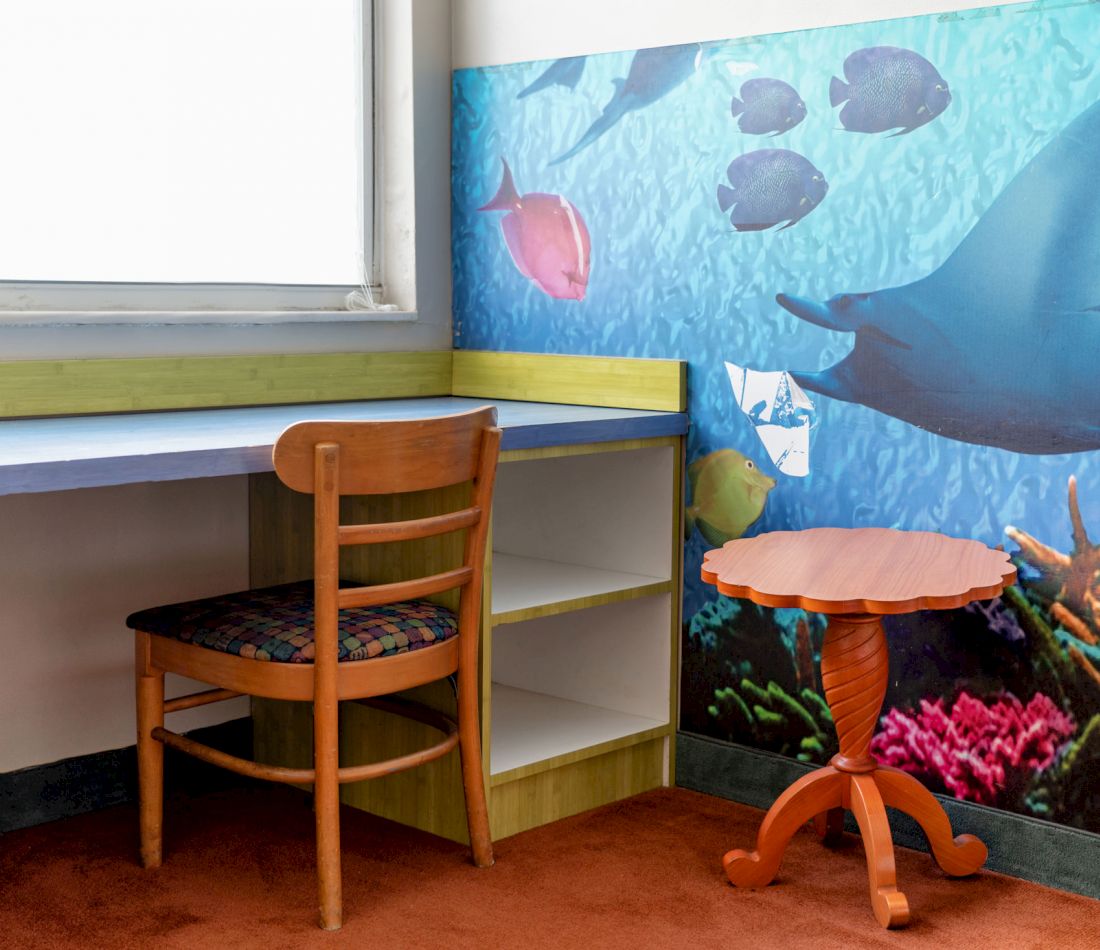 A study area with a desk, chair, table, and bookshelf, featuring an ocean-themed mural.
