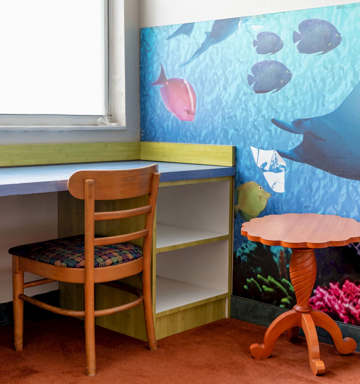 A small room corner with a wooden chair, desk, round table, and an underwater-themed wall mural featuring fish and a ray.