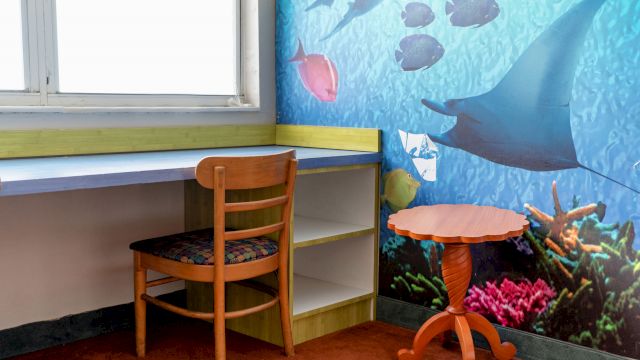 A guest room with a desk, chair, table, and bookshelf in front of a colorful ocean-themed mural.