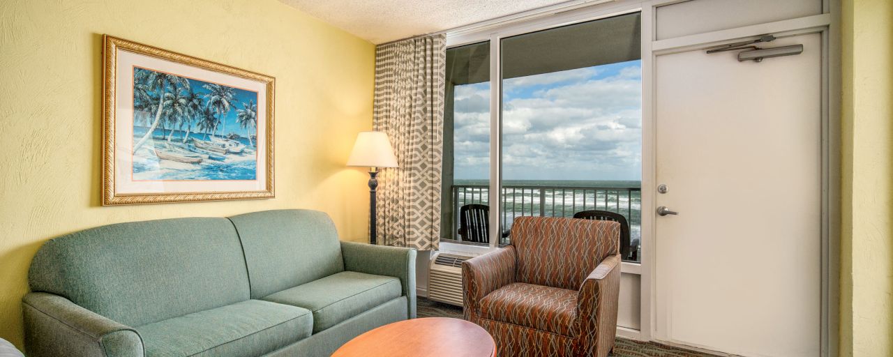 A hotel room with a sofa, chair, coffee table, lamp, and window with a view.