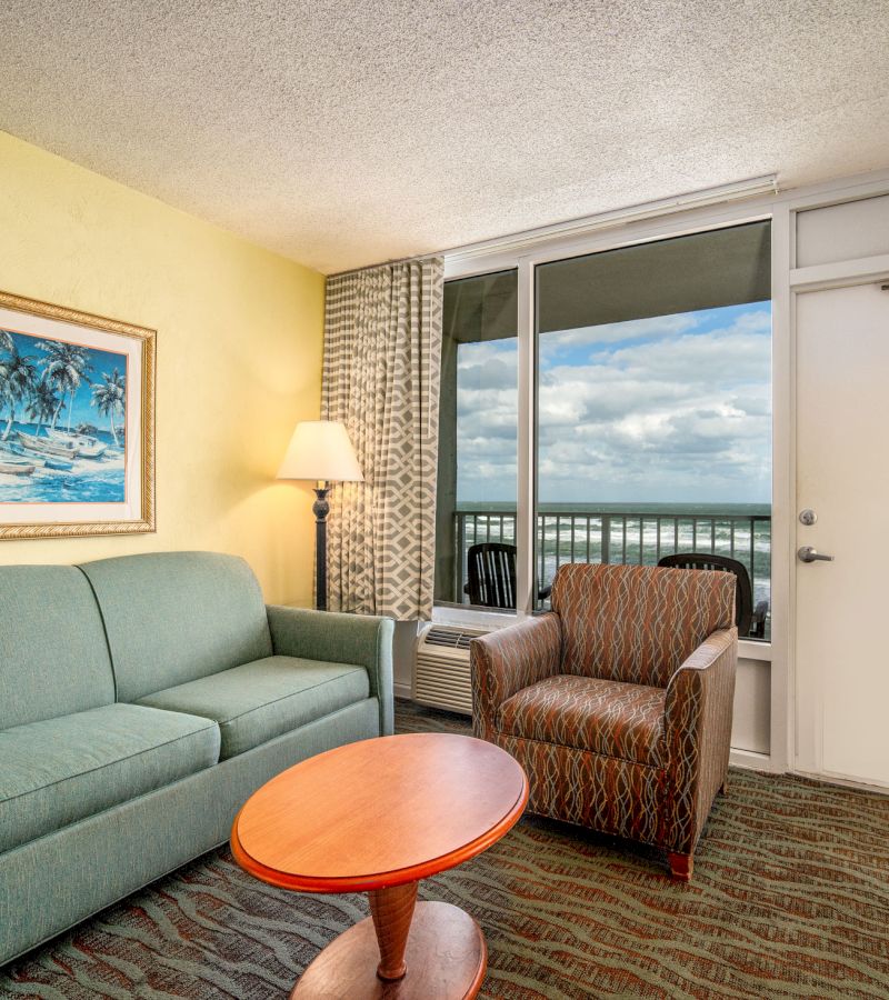 A hotel room living area with a sofa, armchair, coffee table, lamp, and painting, with a view of the ocean through the window.