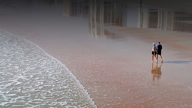 A couple walks along a sandy beach near the shoreline with reflections of buildings on the wet sand in the background.