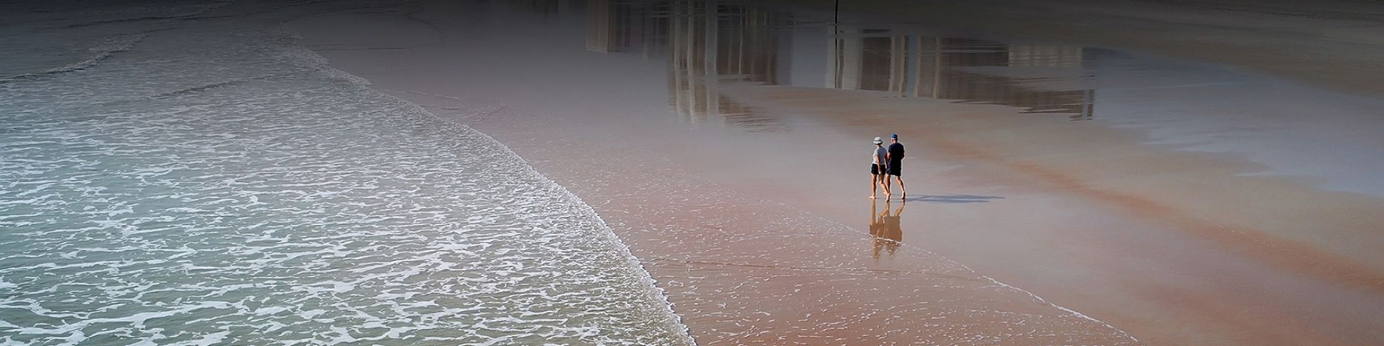 A couple walks along a sandy beach near the shoreline with reflections of buildings on the wet sand in the background.