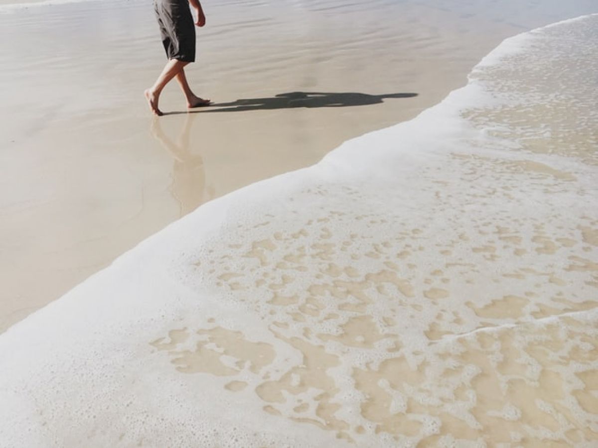 A person walks barefoot along a sandy beach with gentle waves washing up on the shore, creating a serene and peaceful scene.