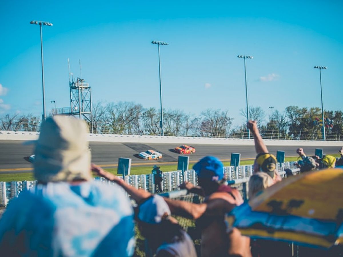 Spectators watch cars racing on a track, with some raising their hands in excitement. The sky is clear, and the atmosphere appears lively.