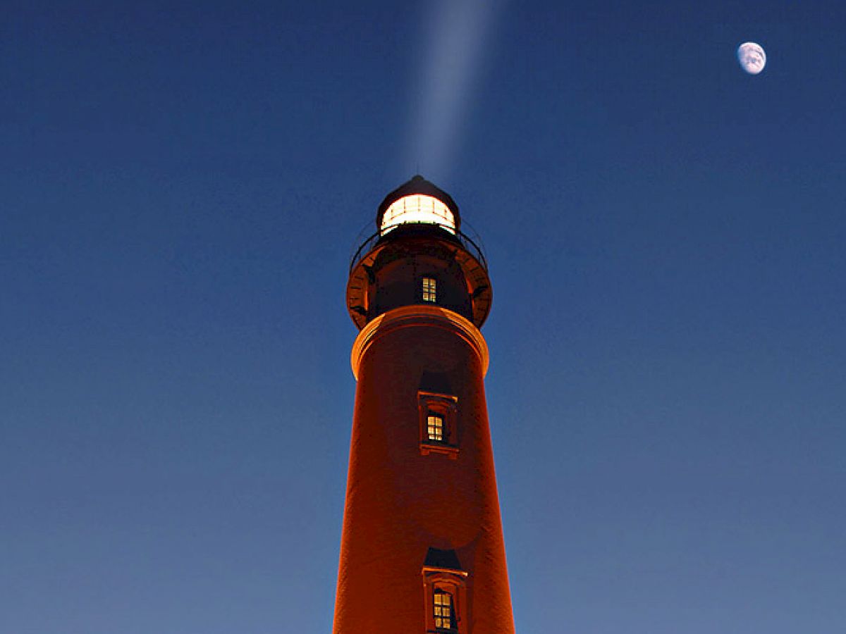 The image shows a lighthouse illuminated at dusk with a light beam shining from the top, and a moon visible in the clear dark blue sky.