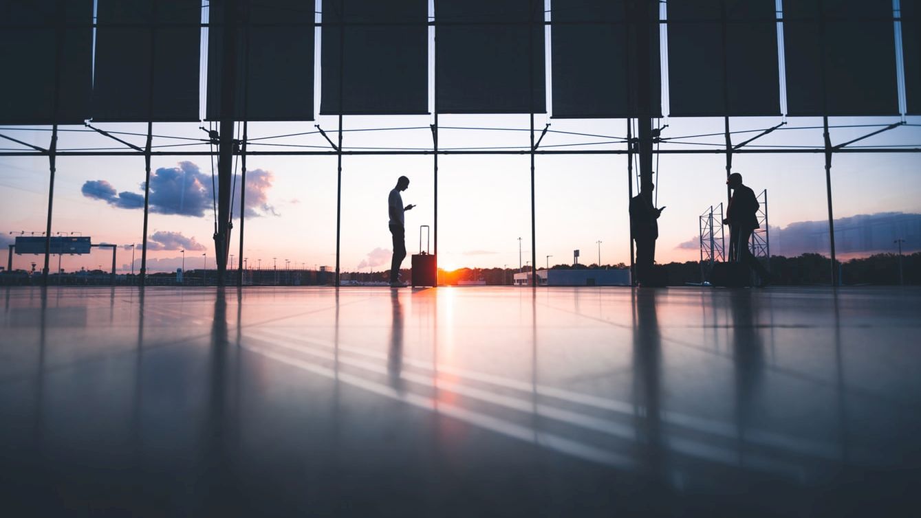 Silhouettes of two people in an airport terminal at sunset, with large windows and a reflective floor. They are standing apart, possibly waiting.