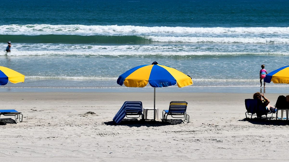 The image shows a beach scene with three blue and yellow umbrellas, lounge chairs and people relaxing while others are in the ocean.