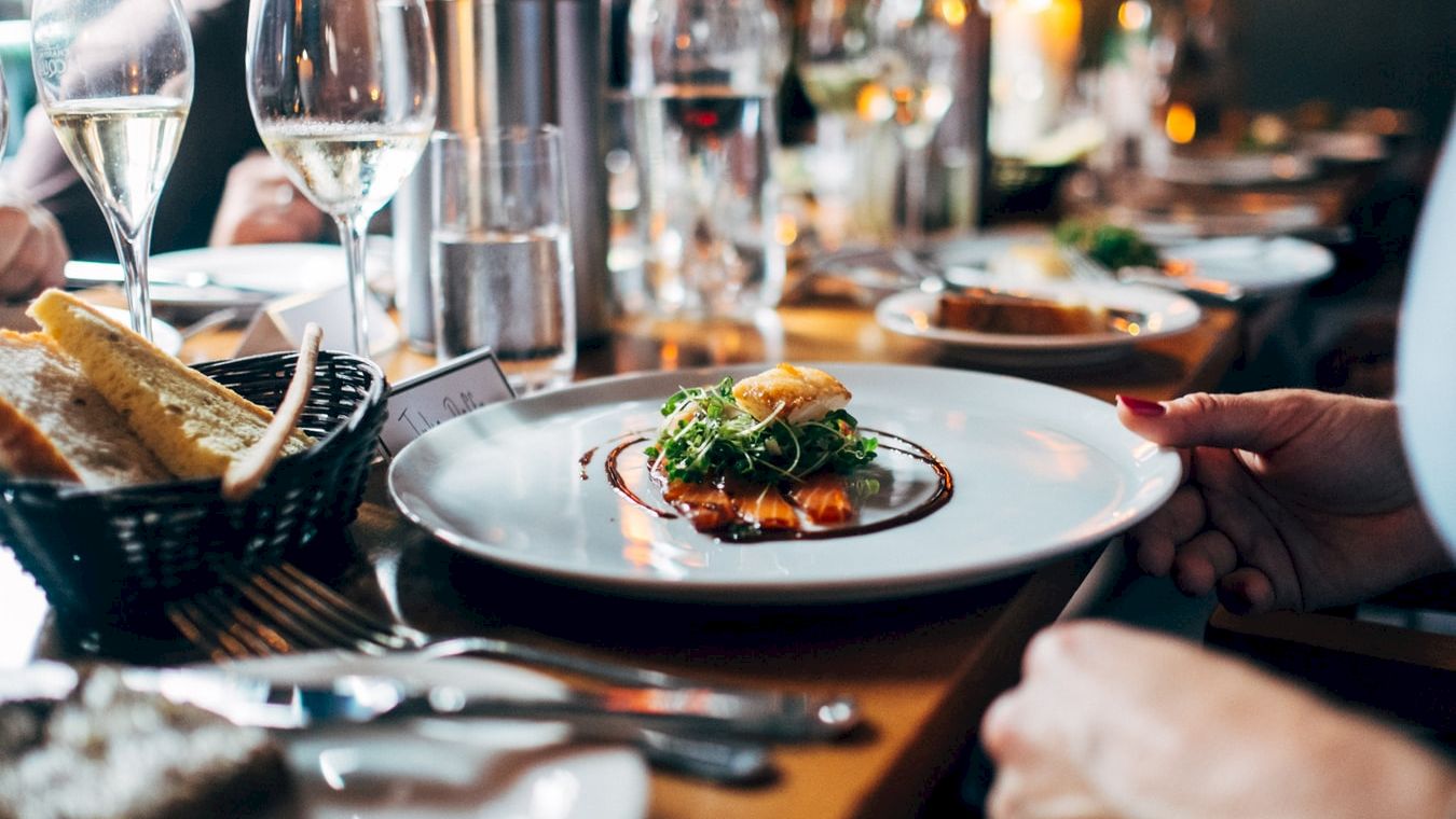 The image shows a dining table setting with plates of food, including a main dish garnished with greens, bread, wine glasses, and cutlery.