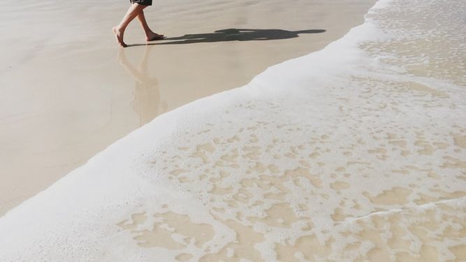 A person walking barefoot on a sandy beach near the shoreline with gentle waves approaching.