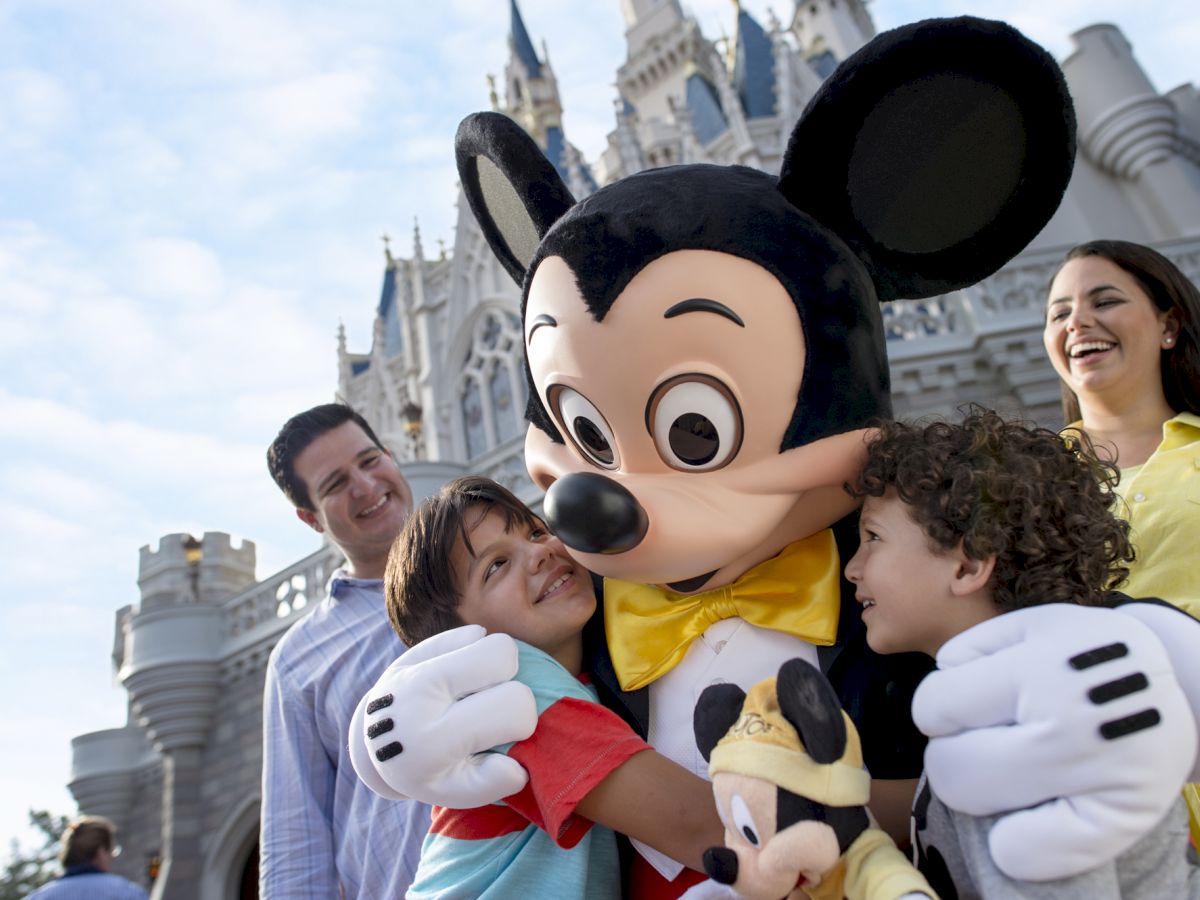 A joyful family meets a large Mickey Mouse character in front of a castle, with children hugging Mickey and everyone smiling in amusement.