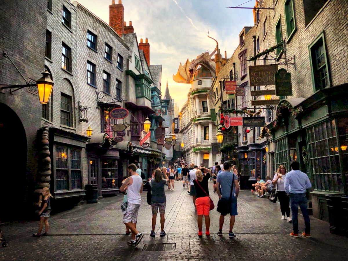 The image shows a bustling alley with shops and people, featuring a dragon atop a distant building, giving a magical, themed experience.