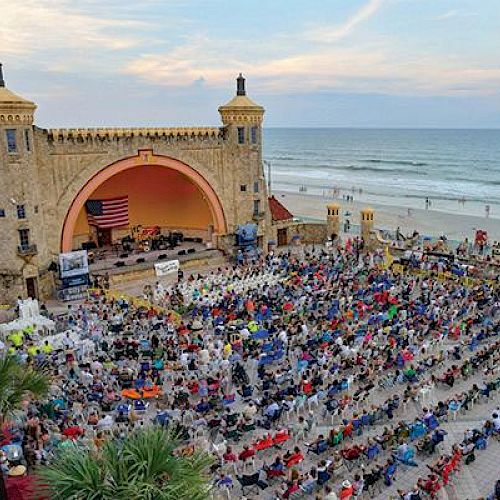 A large crowd gathers in front of a historic open-air venue near a beach, with an American flag displayed on stage under a partly cloudy sky.