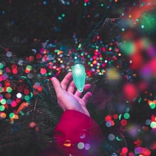 A hand in a red sleeve holds a bright light bulb amidst a background of colorful, blurred lights, creating a festive and vibrant atmosphere.
