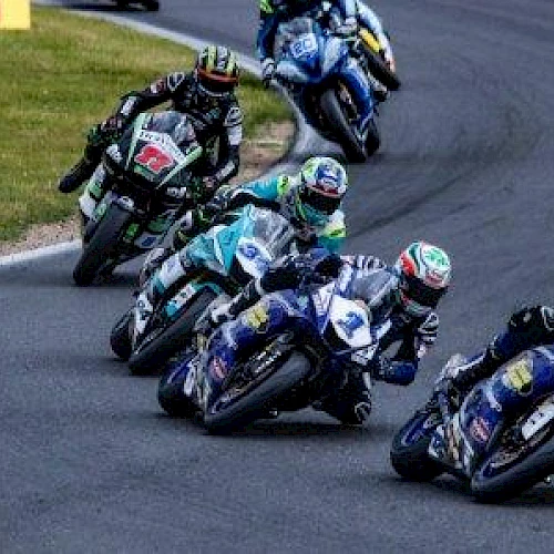 Several motorcyclists are racing on a curved track, leaning into the turn, with a grassy area visible on the left side.