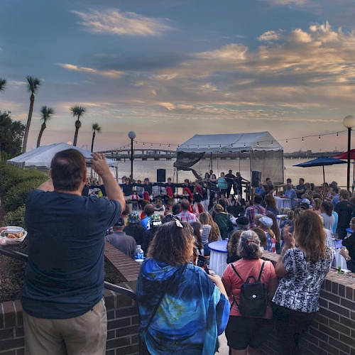 A crowd gathers outdoors by the water under a partly cloudy sky, watching a performance on a stage with tents and palm trees in the background.