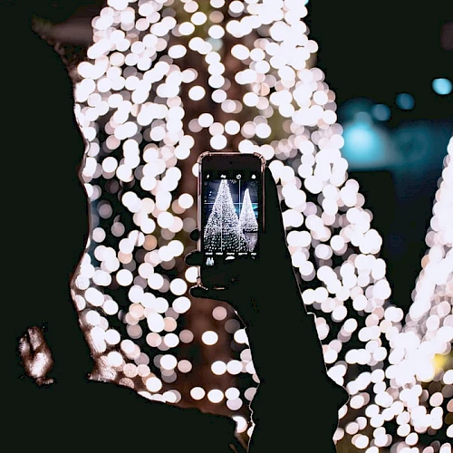 A person holding a smartphone is capturing a picture of illuminated Christmas decorations with blurred lights in the background.