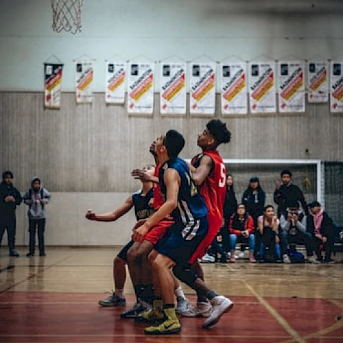 A group of basketball players are in action on the court aiming for the basket, with spectators watching from the background.