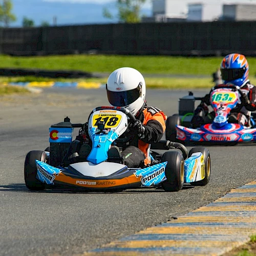 The image shows two people racing go-karts on an outdoor track, with one kart marked #148 leading the other marked #163.