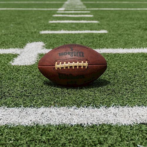 The image shows an American football placed on a football field with green artificial turf and white yard lines.