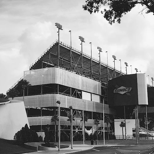 The image depicts a large stadium with multiple levels of seating, light poles, and a prominent entrance. The structure is located near a tree.