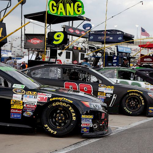 A row of NASCAR race cars lined up in the pit area with crew stations and sponsor logos visible in the background.