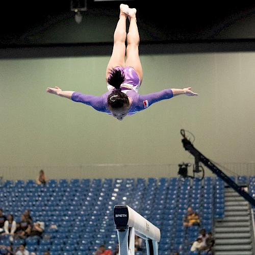 A gymnast in a purple leotard is performing a flip above a balance beam during a competition in an arena with a sparse audience.