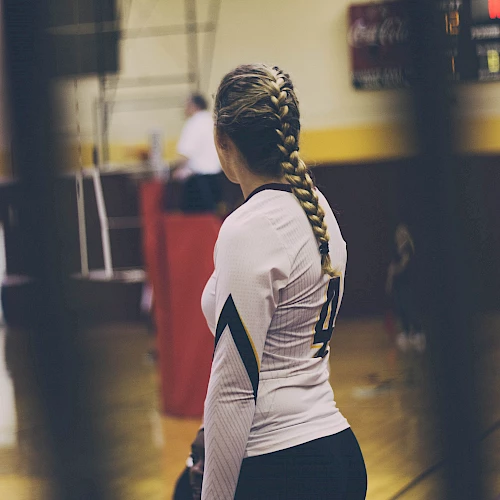A person with a braided hairstyle wearing a volleyball uniform stands in a gymnasium, with equipment and a partial visible figure in the background.