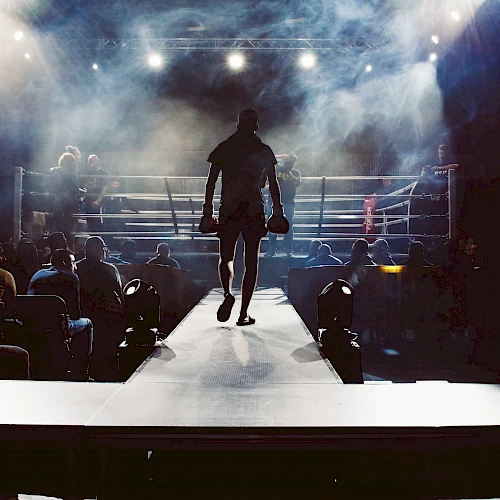 A boxer walks towards the ring under dramatic lighting, with spectators surrounding the area, creating an intense and anticipatory atmosphere.