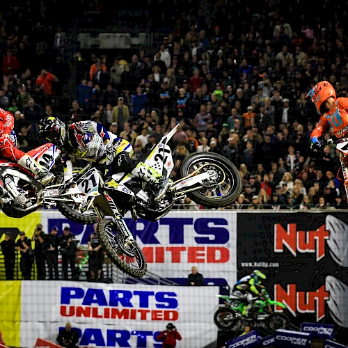 The image shows three motocross riders mid-air during a race, with a large crowd watching in the background and various advertising banners visible.