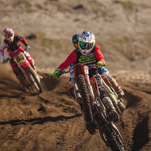 The image shows several motocross riders racing on a dirt track, with the lead rider kicking up dirt while taking a turn.