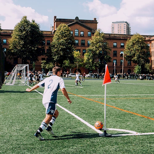 A soccer player is taking a corner kick on a green field, with a red flag marking the corner, and a brick building and trees in the background.