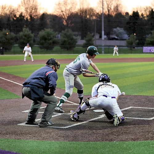 A baseball game in progress, with a batter about to swing, a catcher behind him, an umpire, and fielders in the background.