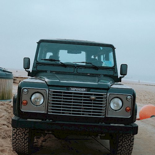 A green off-road vehicle is parked on a sandy beach next to an orange buoy, with a cloudy sky in the background.