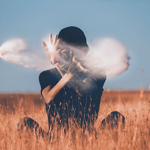 A person sitting in a field with golden grass, obscured by clouds or smoke around their hands, under a clear blue sky in the background.