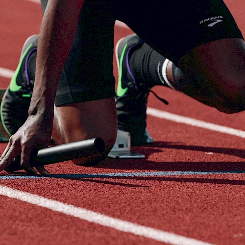 An athlete is on a track, in starting position with a baton, ready for a relay race. The red track and starting blocks are clearly visible.