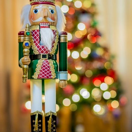 A wooden nutcracker soldier figurine stands in front of a decorated Christmas tree adorned with colorful lights and ornaments, creating a festive atmosphere.