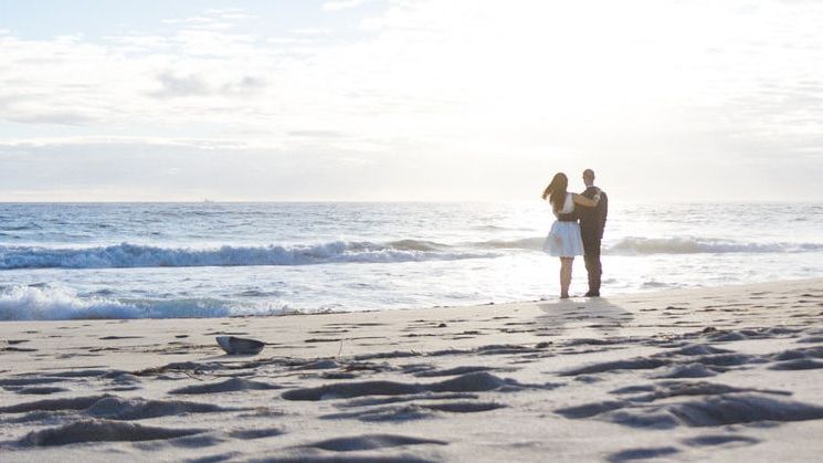 A couple stands on the beach near the shoreline, with scattered seaweed and footprints in the sand, as the sun sets over the ocean.
