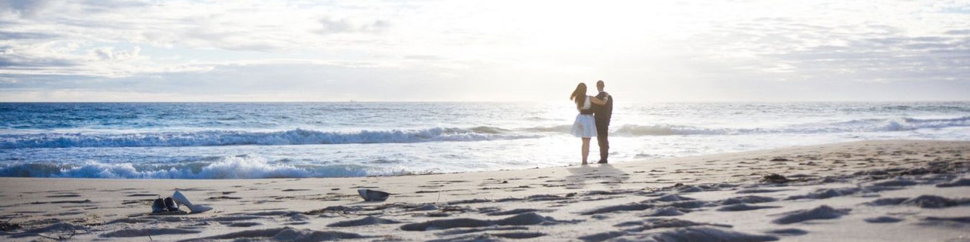 A couple stands on the beach near the shoreline, with scattered seaweed and footprints in the sand, as the sun sets over the ocean.